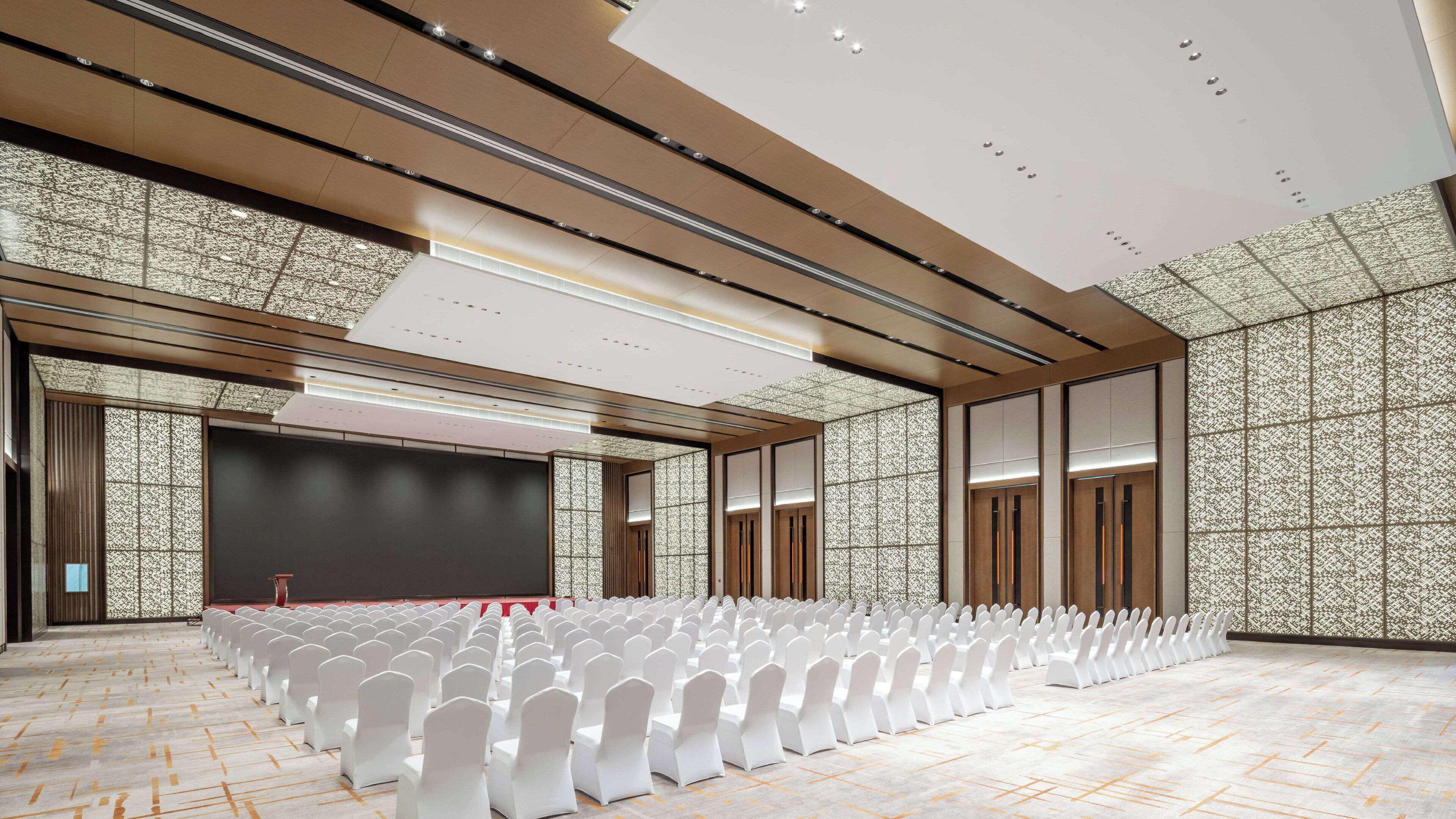 A large conference room with rows of white chairs, a stage, and a patterned wall.