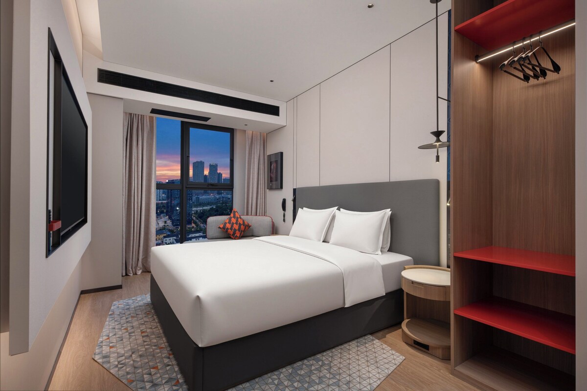 Ein modernes Hotelzimmer mit einem großen Bett, Blick auf die Stadt, einem Kleiderschrank und einem kleinen runden Tisch.