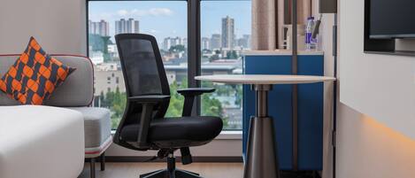 A modern hotel room with a large window, a desk, a chair, a bed, and a TV.