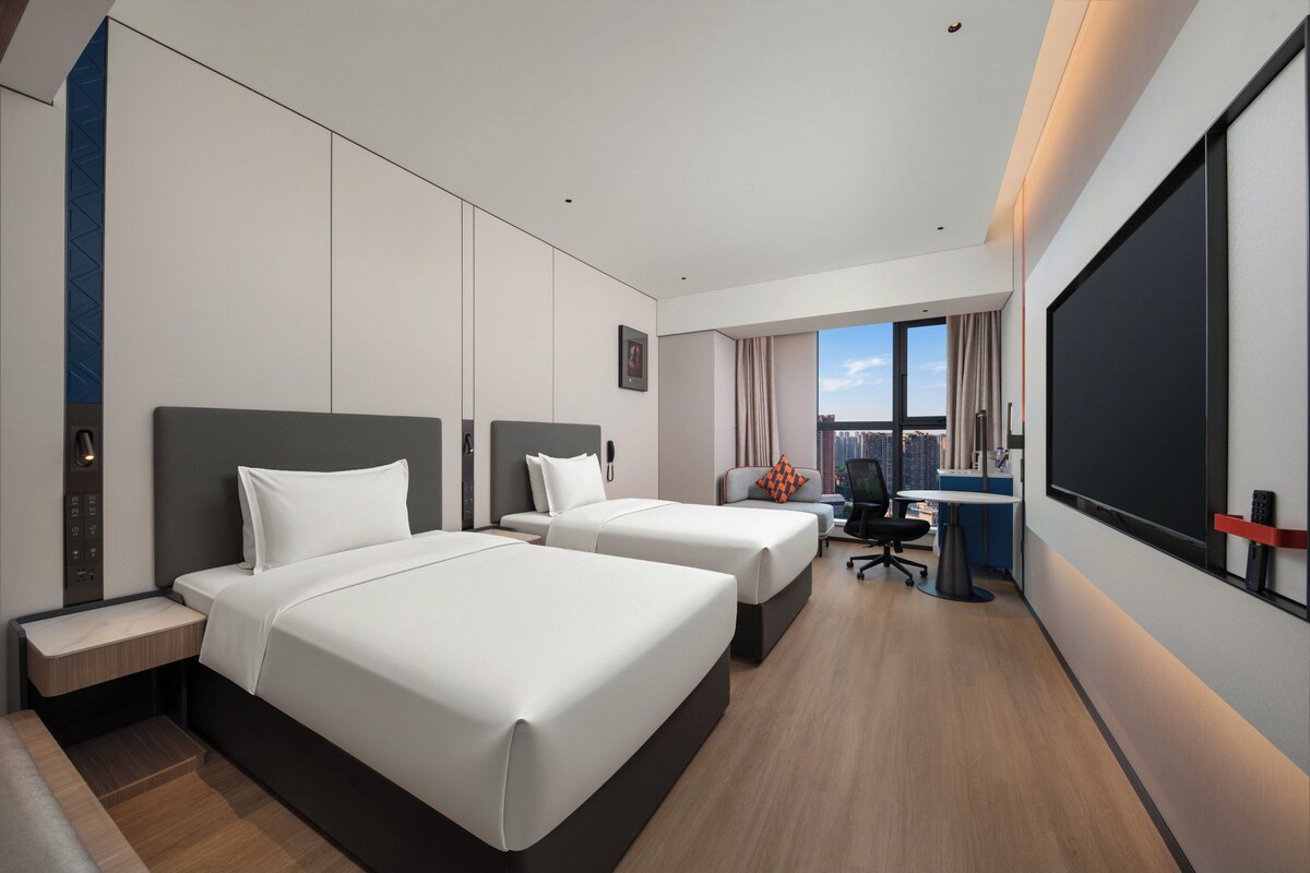 Ein Hotelzimmer mit zwei Betten, einem Schreibtisch, einem Stuhl, einem großen Fenster und Blick auf die Stadt.