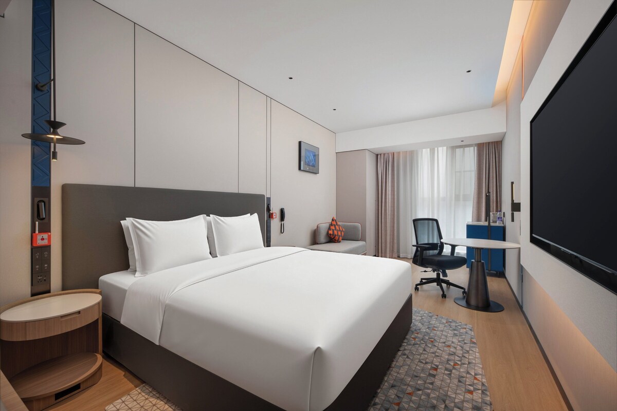 Ein modernes Hotelzimmer mit einem großen Bett, einem Schreibtisch, einem Stuhl, einem Fernseher und einem Fenster mit Vorhängen.