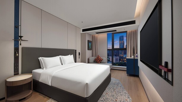 Ein modernes Hotelzimmer mit einem großen Bett, einer Sitzecke, einem großen Fenster mit Stadtblick und einem Flachbildfernseher.