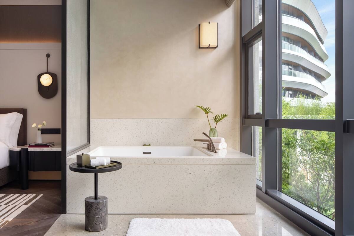 Un dormitorio moderno con una cama grande, una mesita de noche, una bañera y una ventana con vistas a un edificio.