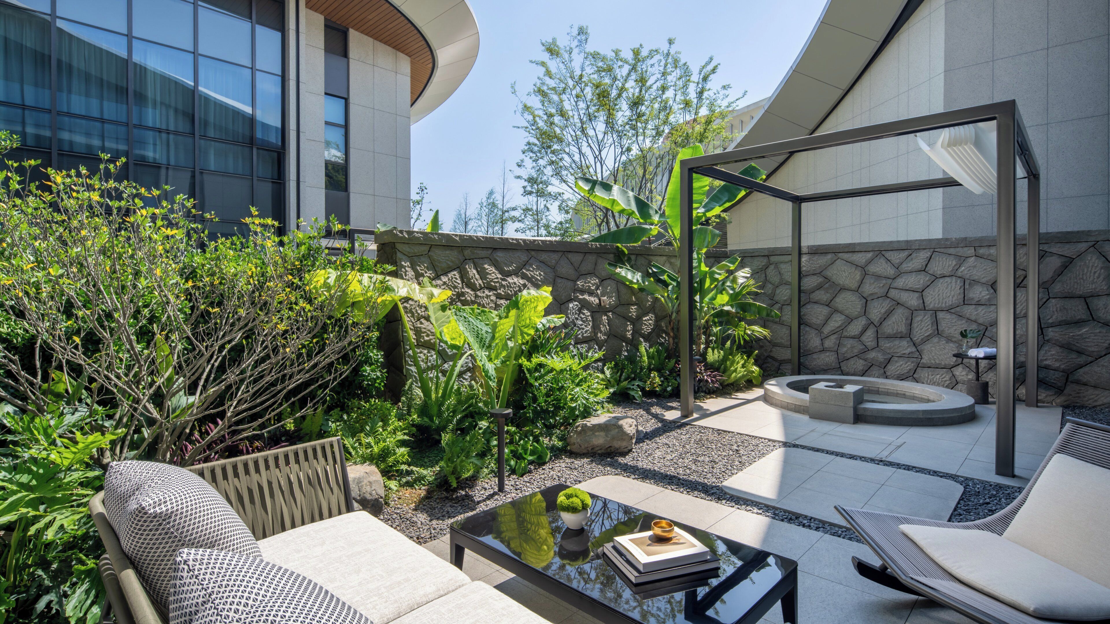 Un patio moderno al aire libre con mobiliario, una mesa de vidrio y un muro de piedra.