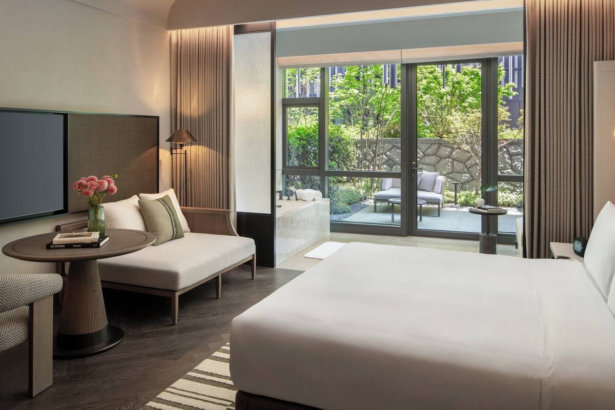 Habitación de hotel con cama, sofá, televisión, una mesita con un jarrón de flores y vistas al exterior.
