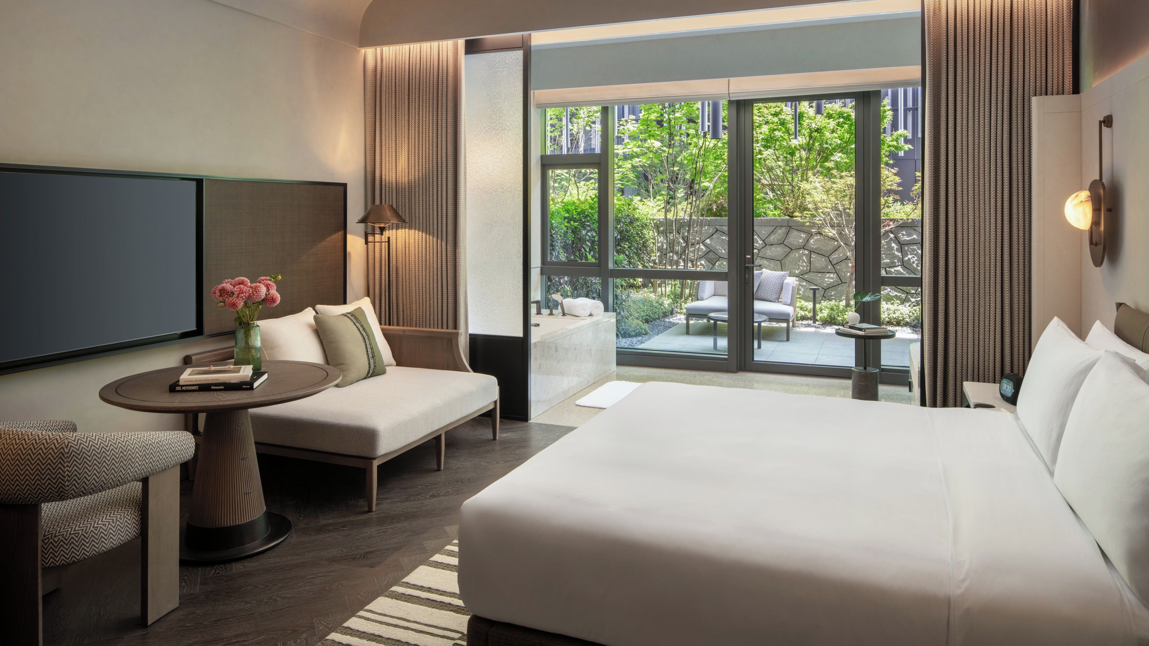 A hotel room with a bed, a sofa, a TV, a small table with a vase of flowers, and a view of the outside.