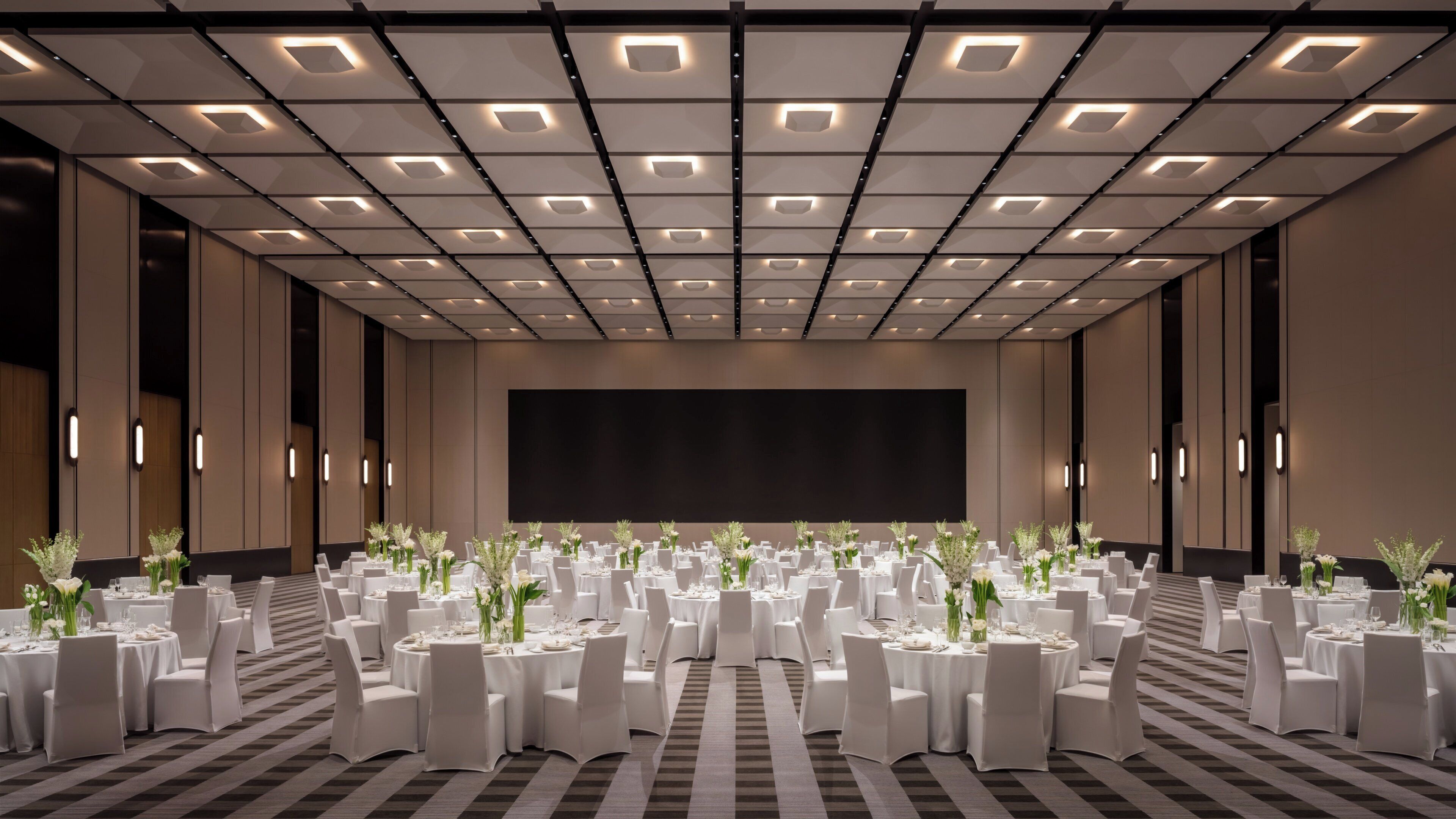 Un amplio salón de conferencias con techo alto, varias mesas dispuestas para eventos y una gran pantalla al frente.