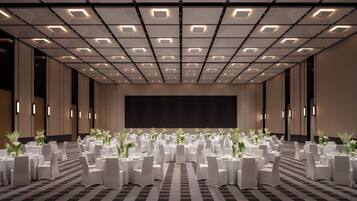 Un amplio salón de conferencias con techo alto, varias mesas dispuestas para eventos y una gran pantalla al frente.