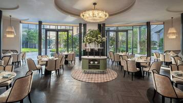 A spacious dining area with round tables, chairs, a central floral arrangement, and large windows.