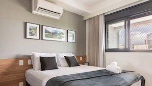 A hotel room with a bed, air conditioning unit, framed pictures on the wall, and a window with curtains. - Expand Pinheiros  (São Paulo)