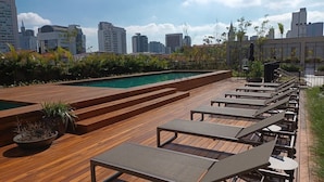 A rooftop pool area with wooden decking, lounge chairs, and a view of the city skyline. - Expand Pinheiros  (São Paulo)