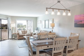A dining area with a wooden table and chairs, a sofa, a lamp, and a painting on the wall. - Point Emerald D-104, Oceanview 3BR 3BA Emerald Isle! (Emerald Isle)