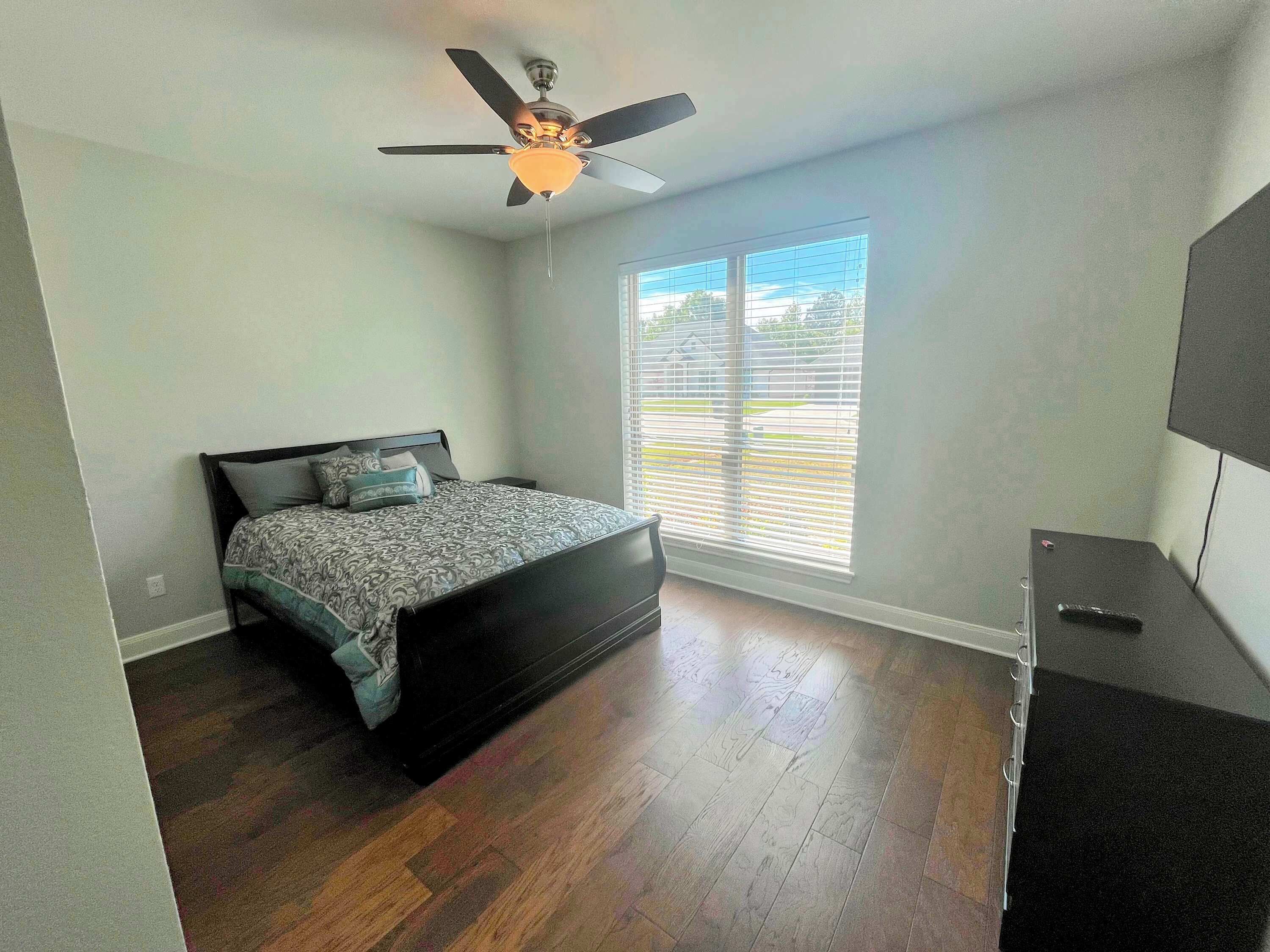 Un dormitorio con cama, ventilador de techo, ventana con persianas y un mueble para televisor.