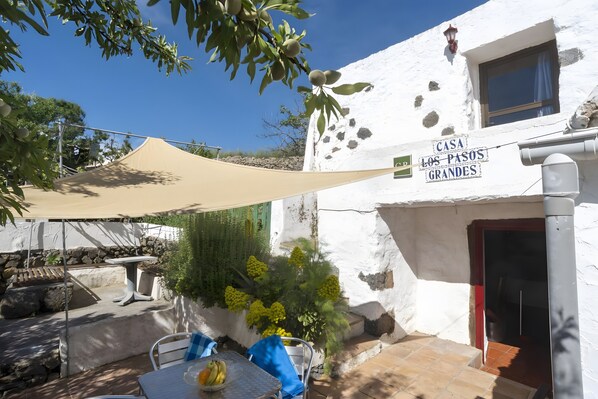 Un bâtiment blanc avec une enseigne indiquant « CASA LOS PASOS GRANDES », une terrasse avec une table et des chaises, et une grande voile d’ombrage.