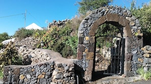 Un arc en pierre surmonté d’un portail en métal, entouré de végétation et adossé à une colline sur laquelle se trouve une structure blanche en arrière-plan.