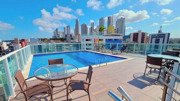 Ein Pool auf dem Dach mit Glasgeländern, Korbstühlen und einer Stadtkulisse im Hintergrund.