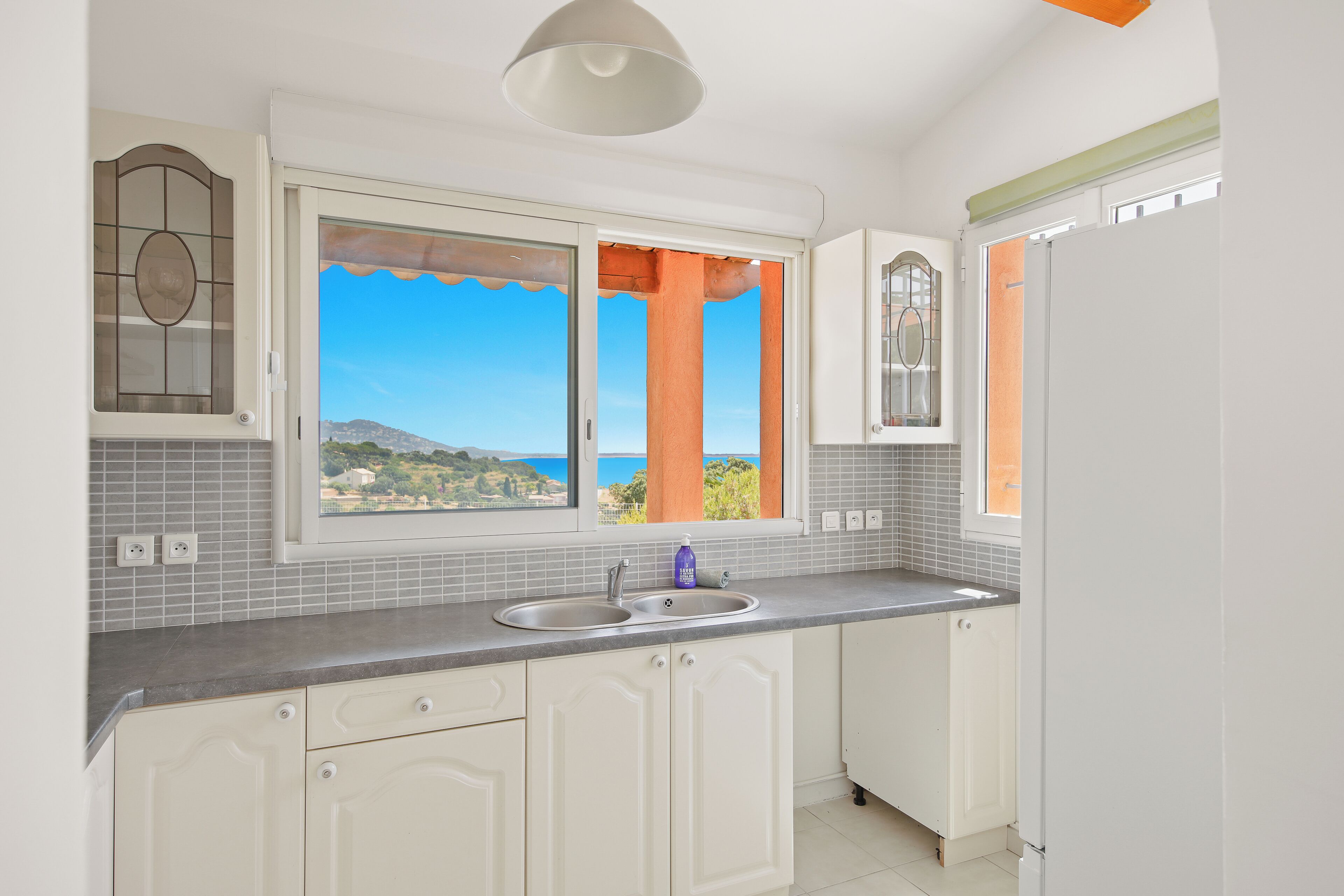 A kitchen with a view of the sea, a window with orange curtains, and a white refrigerator.