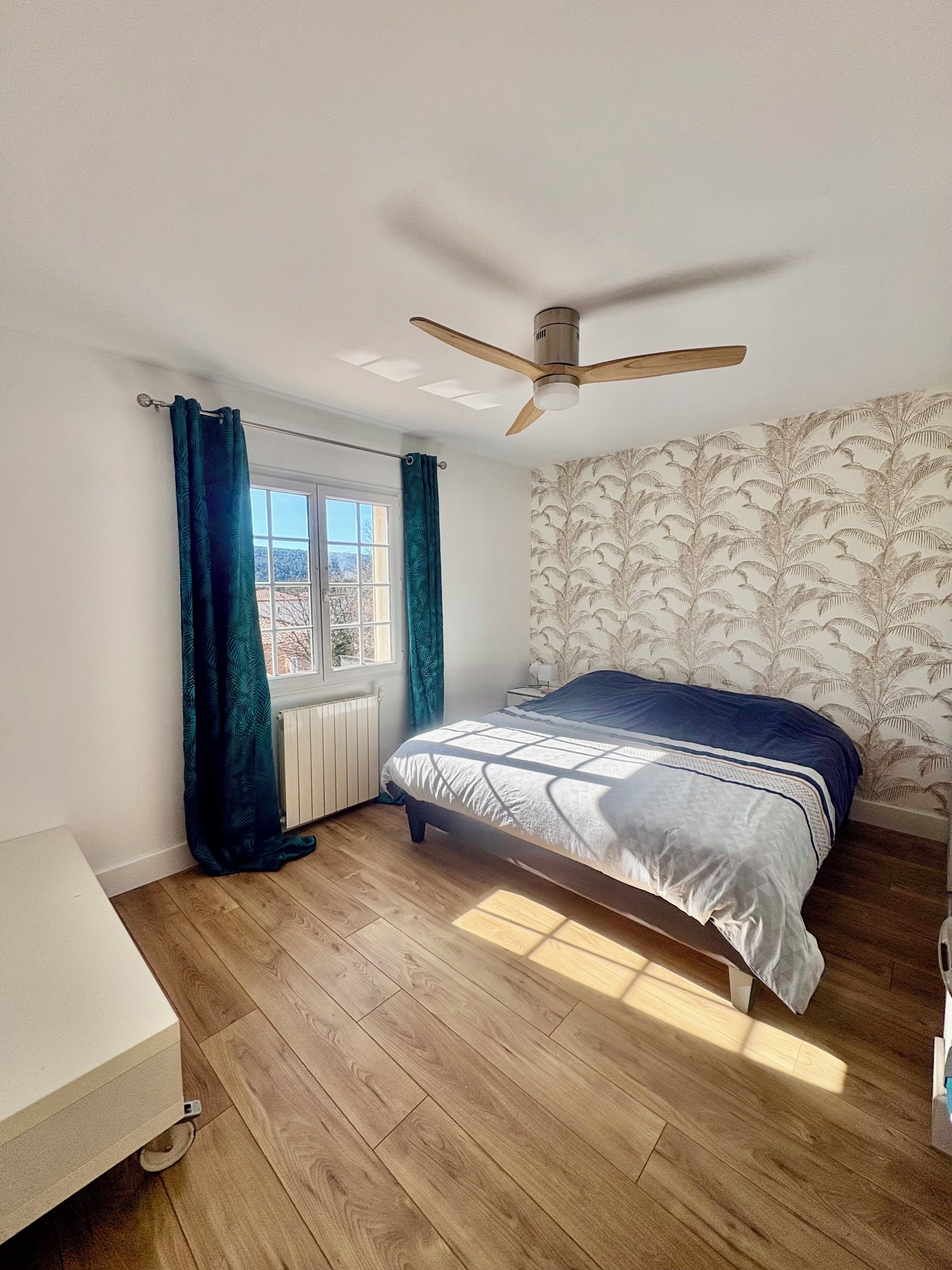 A bedroom with a bed, a ceiling fan, a window with curtains, a radiator, and a wooden floor.