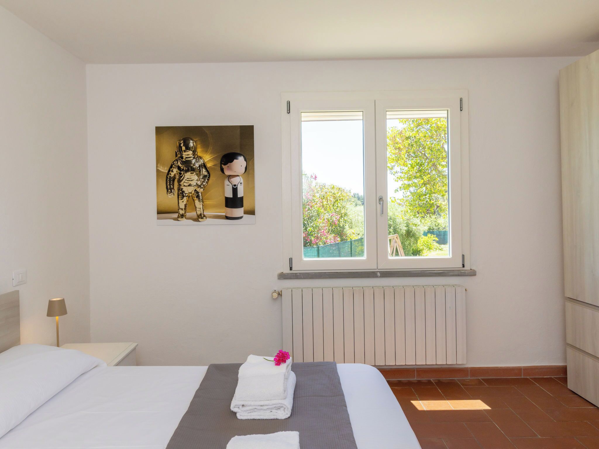 Une chambre d’hôtel avec un lit, un radiateur, une fenêtre donnant sur un espace verdoyant et un tableau accroché au mur.