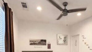 Una camera da letto con un letto, una ventola a soffitto, una finestra con tende, un quadro appeso al muro e un piccolo comodino.
