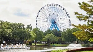 Una ruota panoramica con la scritta 'Texas', circondata da alberi e un lago con una fontana.