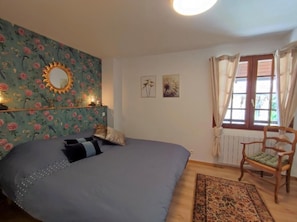 A bedroom with a bed, a wooden chair, a window with curtains, and floral wallpaper. - Holiday Home 'Gite Les Rosiers' with Private Garden (Eu)