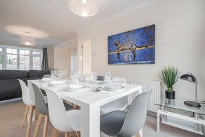 A modern dining area with a white table set for four, grey chairs, a glass-top table, and a large abstract painting on the wall. - Charming 3 Bed Hse Family and Long Stay (Maidenhead)