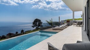 Ein moderner Poolbereich mit freiem Blick auf das Meer, einer Holzterrasse und einer Lounge mit Sesseln und einem Tisch.