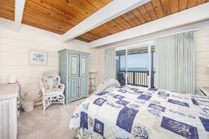 Ein Schlafzimmer mit Holzbalkendecke, einem großen Bett mit Quilttdecke, einem Korbsessel, einem Schrank und Meerblick durch eine Schiebetür.