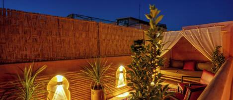 A rooftop terrace with a wooden wall, potted plants, a seating area with cushions, and a decorative lamp.