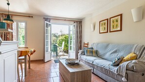 Una sala de estar con un sofá, una mesa de centro, una mesa de comedor con sillas y vistas al exterior por una puerta abierta.