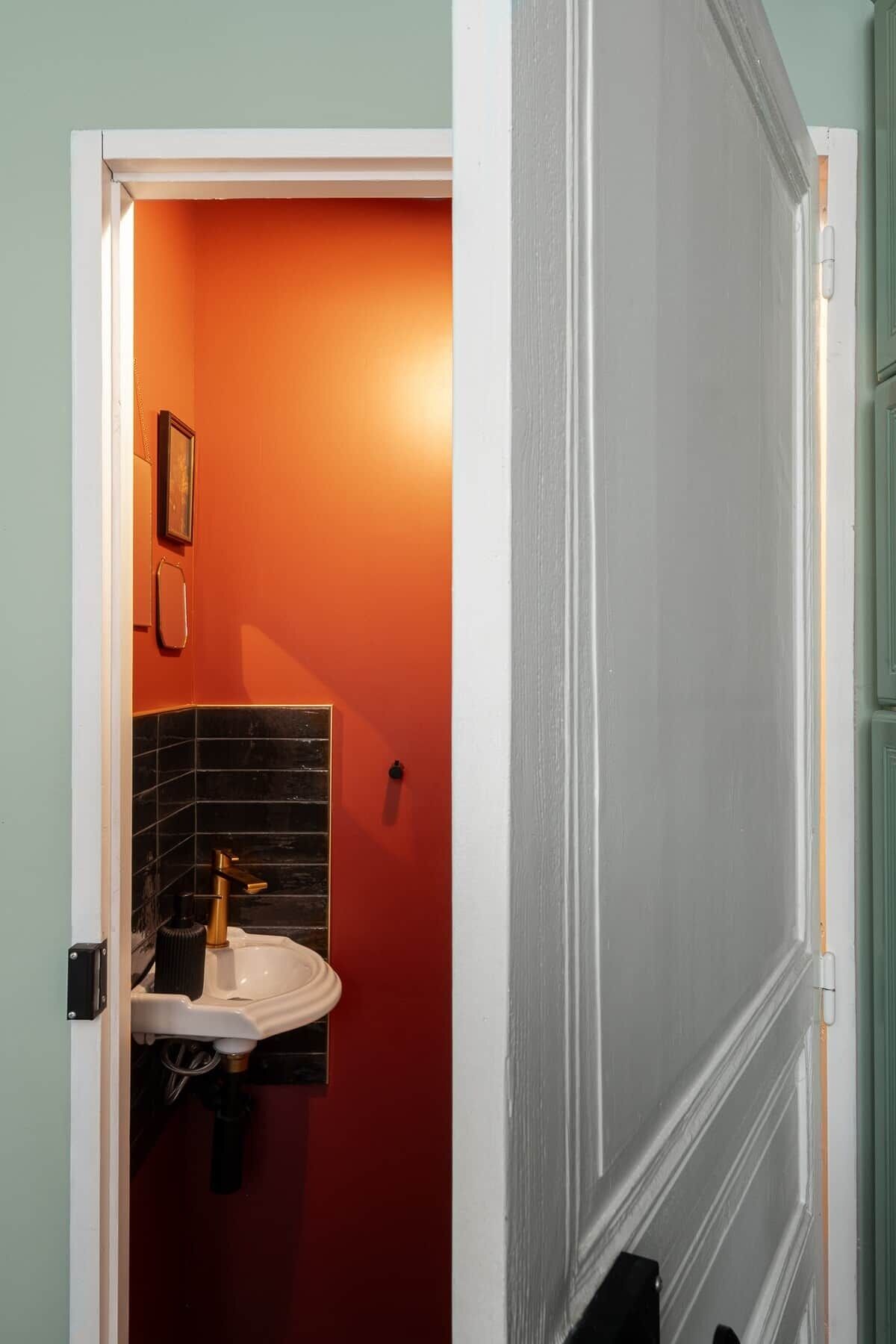 Casa de banho com porta branca, parede laranja, painel de azulejos pretos e lavatório branco com torneira dourada.