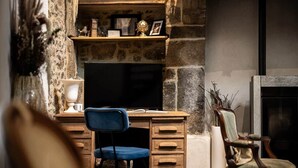 A stone fireplace, a television on a wooden desk, a blue chair, and a wooden chair with a cushion. - LE COTTAGE - Charming gîte at the foot of the Château (Combourg)