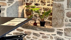 Um muro de pedra com uma abertura em arco, uma mesa com cadeiras e plantas em vasos.