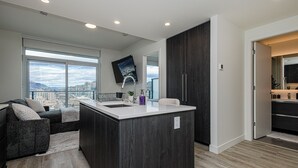 A modern kitchen with a large island, a dining area with a sofa, and a view of a cityscape. - Modern 1-bedroom condo, Lakeview, sleeps 4,  pool, gym, amazing amenities. (Kelowna)