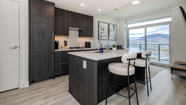 A modern kitchen with dark wood cabinets, a central island with bar stools, and a large window offering a scenic view. - Modern 1-bedroom condo, Lakeview, sleeps 4,  pool, gym, amazing amenities. (Kelowna)