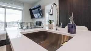 A modern kitchen with a stainless steel sink, a high-arc faucet, a mounted television, and a city view. - Modern 1-bedroom condo, Lakeview, sleeps 4,  pool, gym, amazing amenities. (Kelowna)