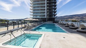 A rooftop pool with a view of a high-rise building, lounge chairs, and a clear sky. - Modern 1-bedroom condo, Lakeview, sleeps 4,  pool, gym, amazing amenities. (Kelowna)