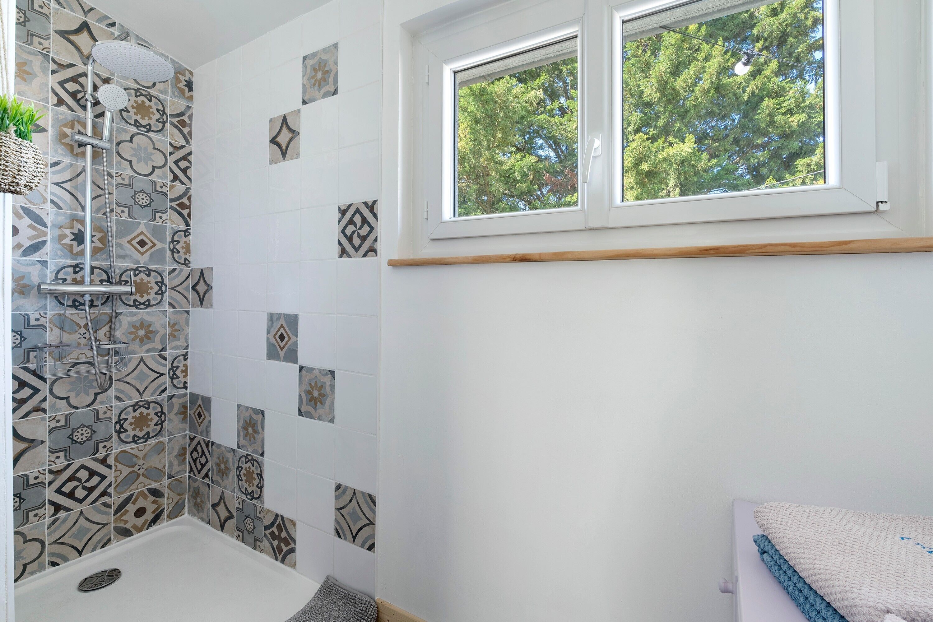 Une salle de bain avec des carreaux décorés, une douche, une fenêtre donnant sur les arbres et un porte-serviettes.