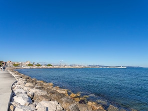 A coastal walkway with a rocky shoreline, a lighthouse in the distance, and buildings along the coast. - Port de La Gavine by Interhome (Hyères)