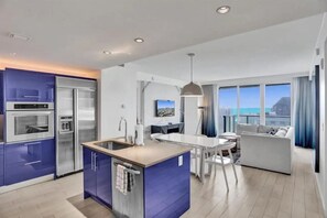 Eine moderne Küche mit integrierten Geräten, ein Essbereich mit weißem Tisch und Stühlen sowie ein Wohnzimmer mit Sofa und großem Stadtblick.