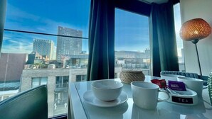 Ein Zimmer mit Tisch, gedecktem Tee-Service, einer Lampe und Blick auf Stadtgebäude.