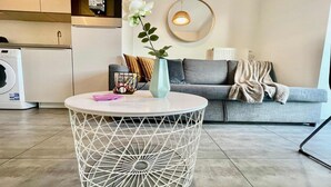 Ein Wohnzimmer mit einem runden weißen Tisch, einem grauen Sofa, einer Blumenvase und einer Waschmaschine im Hintergrund.