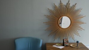 A room with a blue armchair, a wooden cabinet with drawers, and a sunburst mirror on the wall.