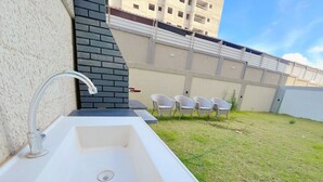 A white outdoor sink with a modern faucet, a grassy area with white chairs, and a building with a brick wall and windows in the background. - Jardim dos Corais - Miramar (João Pessoa)