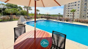 A swimming pool with a wooden deck, outdoor seating, and a view of a residential building in the background. - Jardim dos Corais - Miramar (João Pessoa)