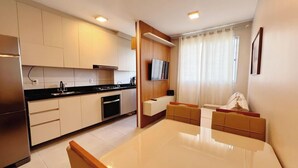 A modern kitchen with white cabinets, a black countertop, and a built-in oven. There is a dining area with a white table and a sofa. - Jardim dos Corais - Miramar (João Pessoa)