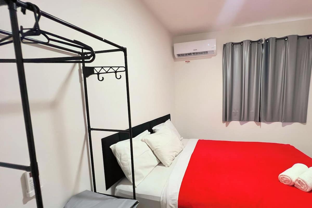 A single bed with a red bedspread, a black metal wardrobe, a grey curtain, and an air conditioning unit on the wall.