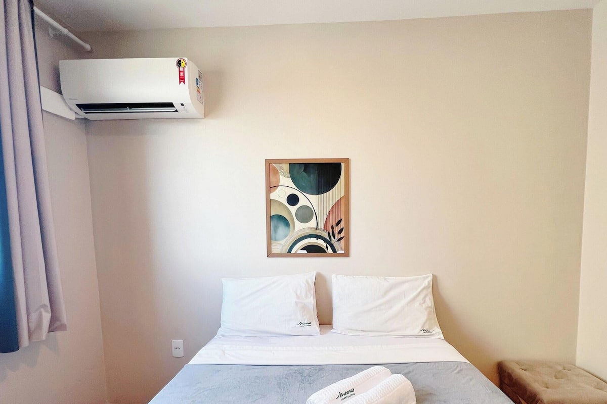 A hotel room with a bed, white pillows, a wall-mounted air conditioner, and a framed abstract painting.