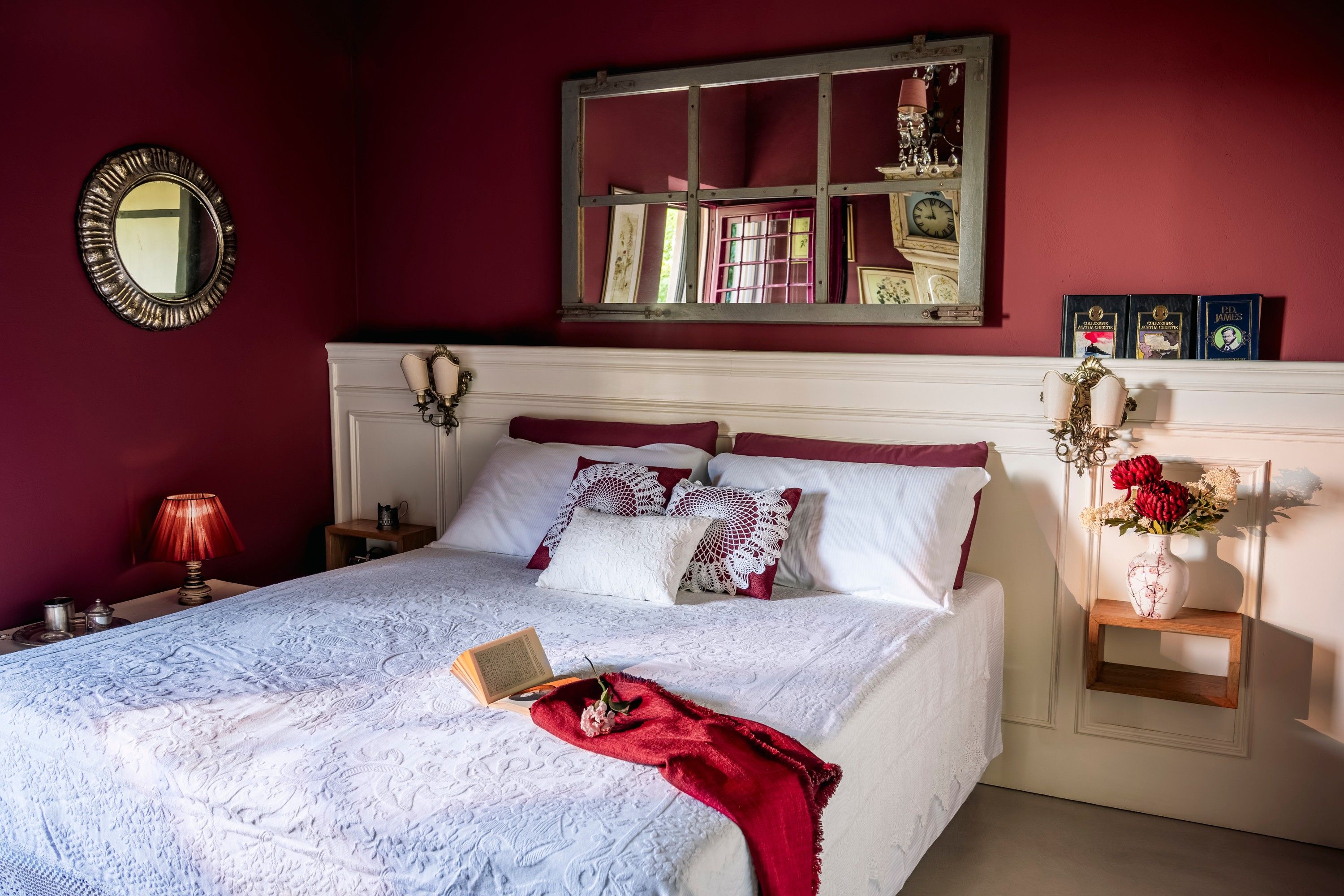 A neatly made bed with a white bedspread and red throw, a bedside table with a lamp, and a wall-mounted mirror.